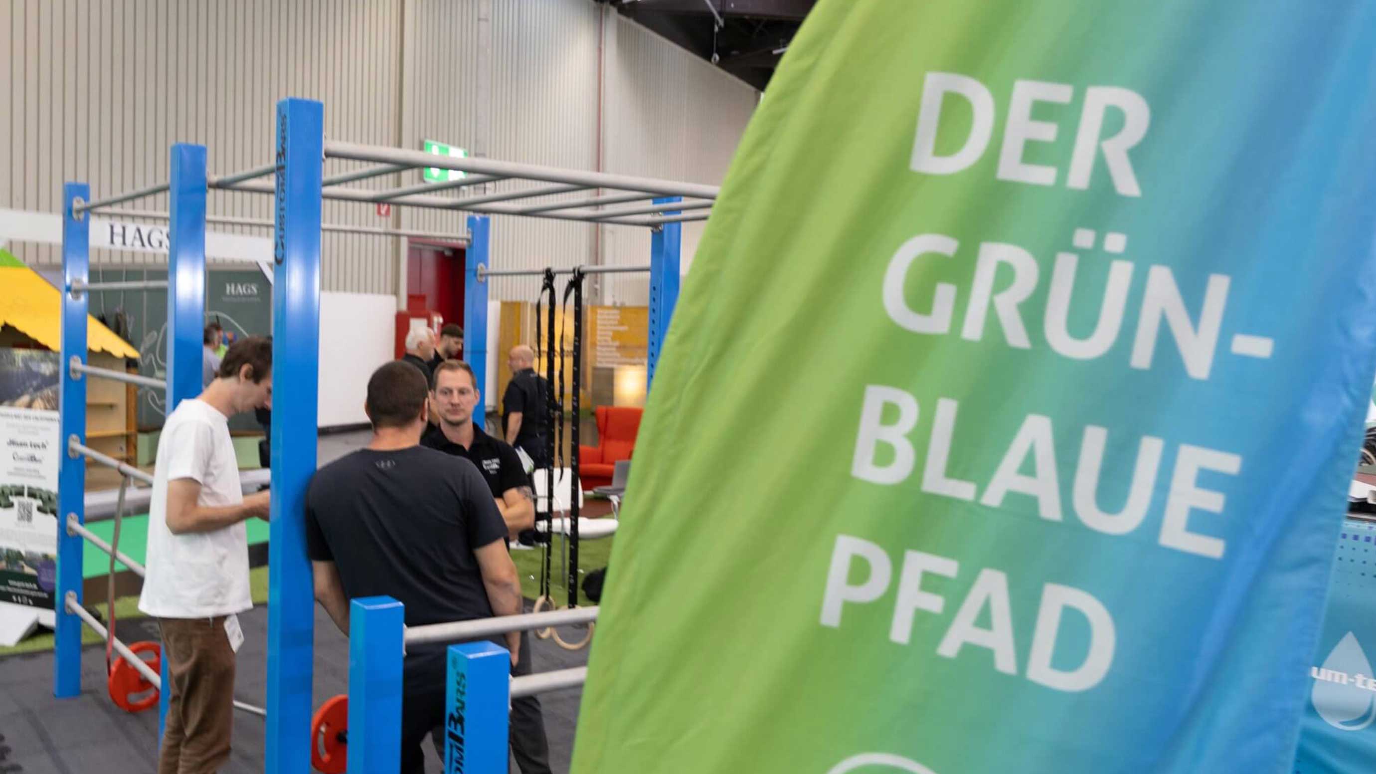 Messebesucher vor blauen Fitnessgeräten; vorn Beachflag "Der Grün‑Blaue Pfad"