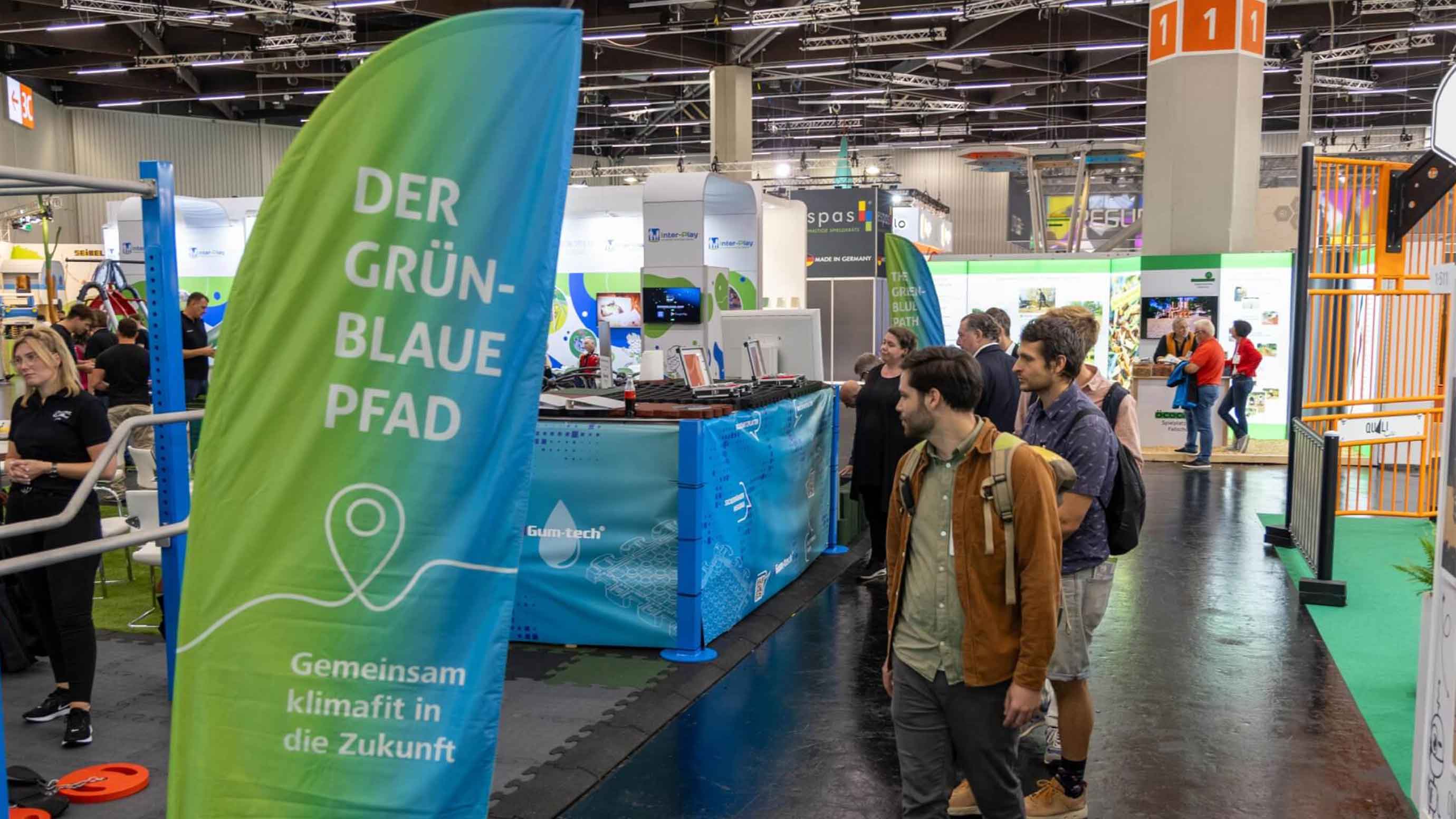 Besucher gehen durch eine Messehalle mit Banner Der Grün‑Blaue Pfad und Ständen.